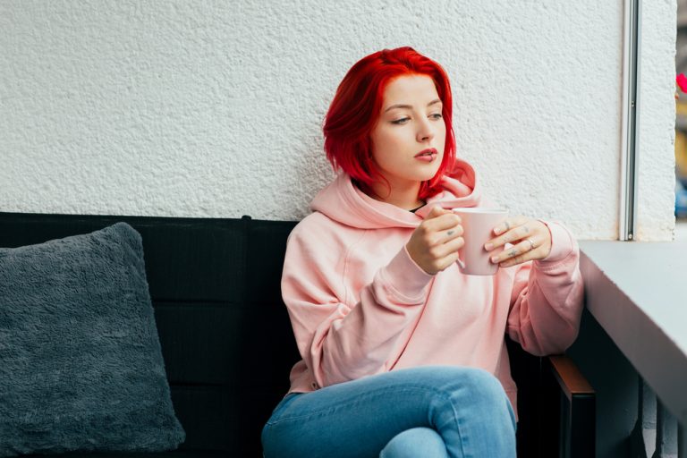 Eine rothaarige traurige Frau in einem rosa Sweatshirt sitzt und trinkt Tee