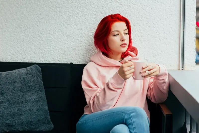 Eine rothaarige traurige Frau in einem rosa Sweatshirt sitzt und trinkt Tee