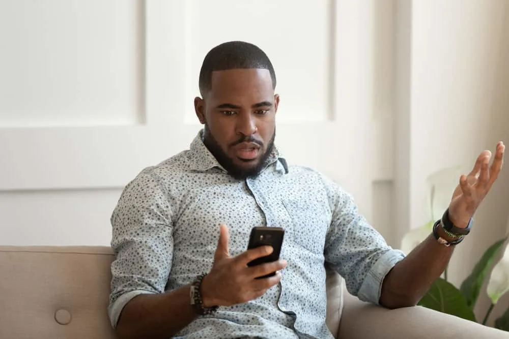 Ein schwarzer Mann in einem Hemd sitzt auf der Couch und schaut verwirrt auf sein Handy