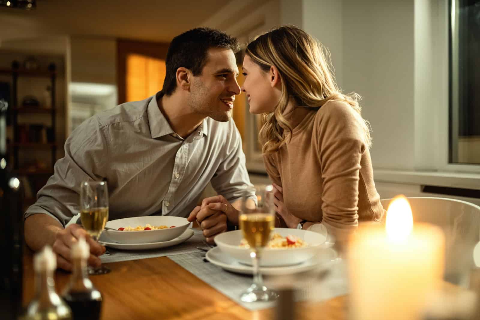 Romantisches Paar, das sich beim Abendessen am Esstisch küssen will, während es Händchen hält.