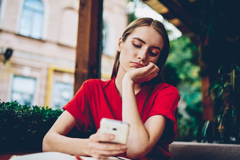 nachdenkliche Frau, die rotes T-Shirt trägt und ihr Telefon im Café betrachtet