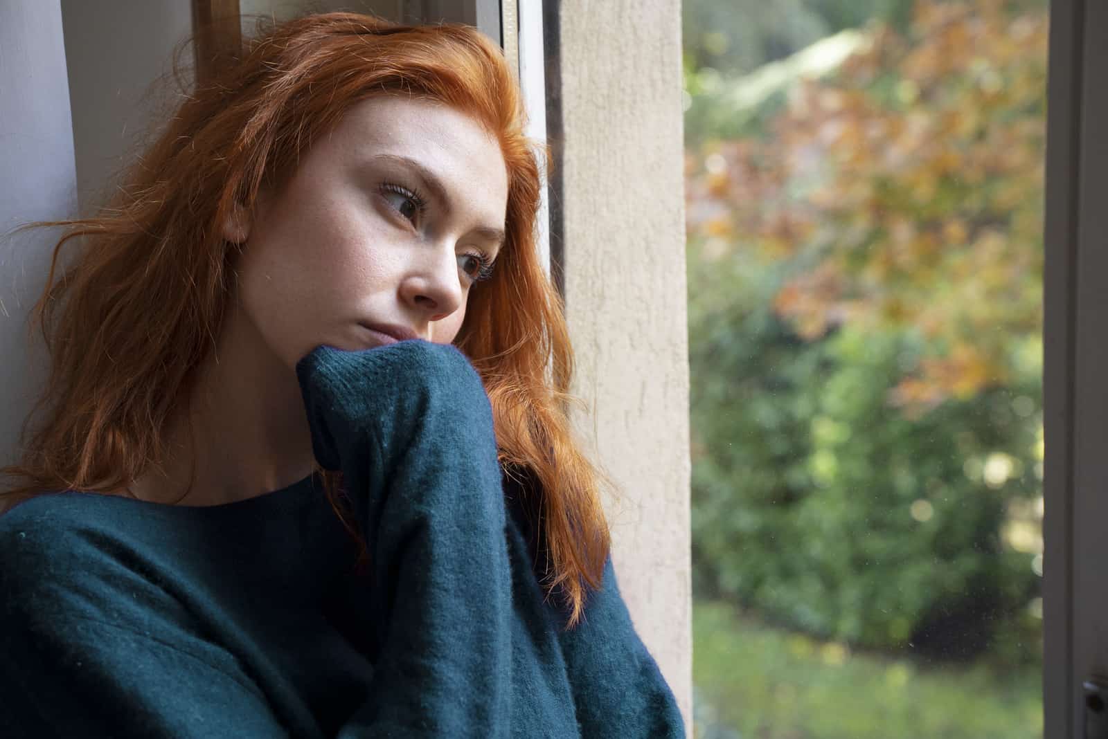 traurige rothaarige Frau, die gedankenverloren durch das Fenster schaut