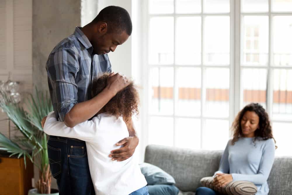 Das Mädchen umarmt ihren Vater vor einer traurig sitzenden Mutter