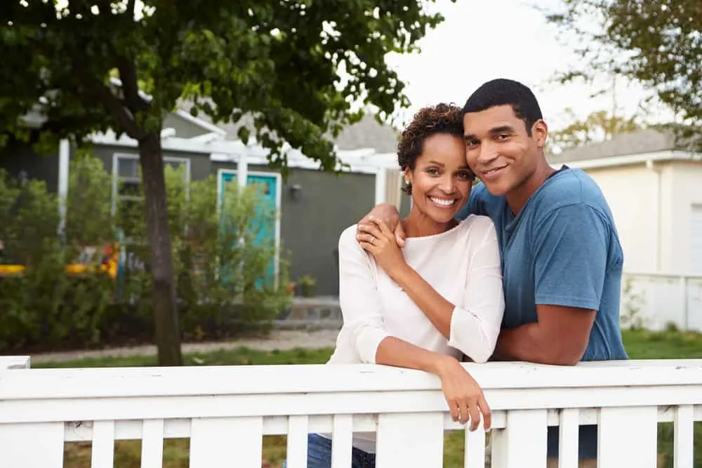 Draußen, neben einem weißen Zaun, umarmen sich ein schwarzer Mann und eine schwarze Frau