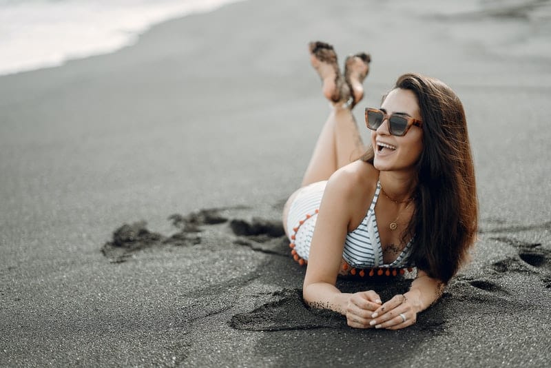 Eine lächelnde Frau in einem weißen Kleid mit Sonnenschirmen liegt an einem Sandstrand