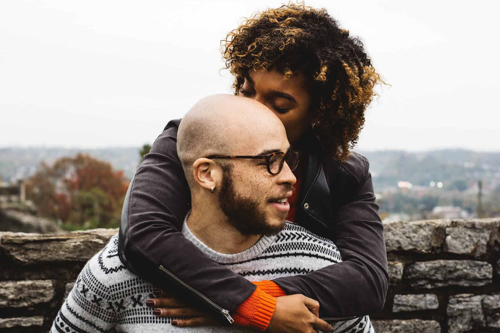 Eine schwarze Frau mit krausem Haar hinter dem Rücken umarmt einen kahlen Mann mit Brille