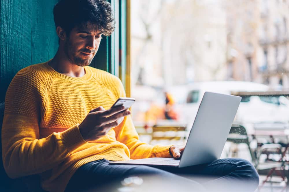 Ein Mann in einem gelben Pullover mit einem Laptop auf dem Schoß schreibt eine SMS auf sein Handy
