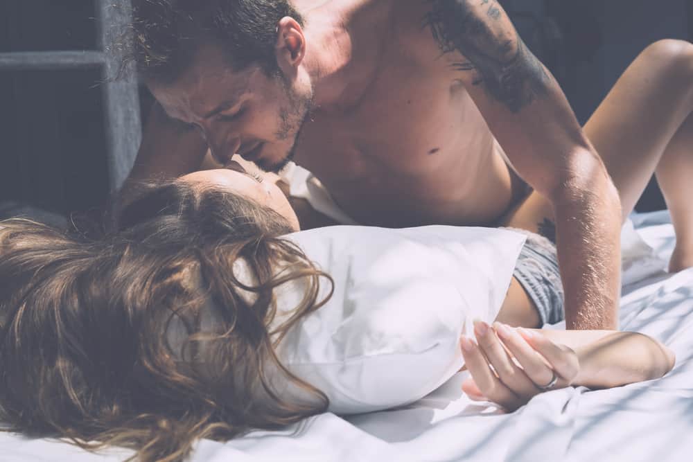 Ein Mann mit einem Tattoo auf der Schulter küsst eine halbnackte Frau auf dem Bett