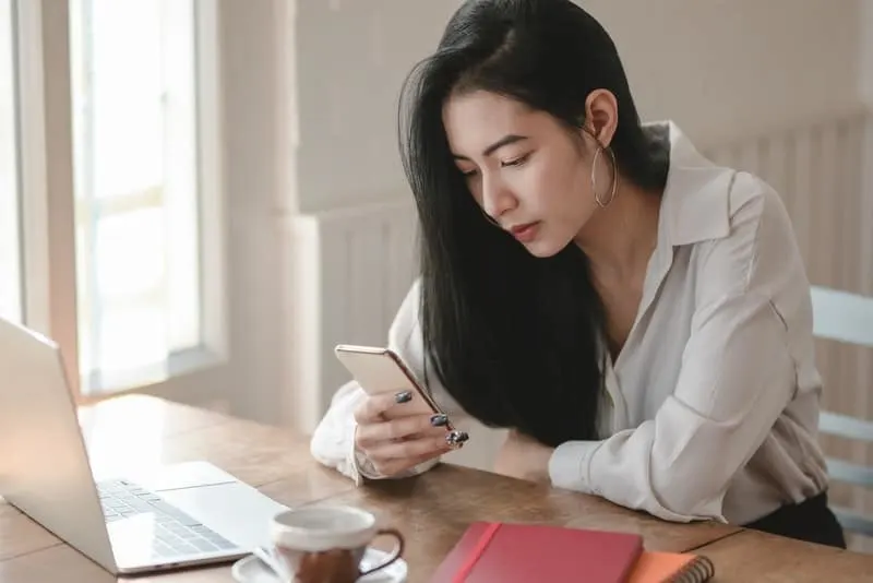 Eine besorgte asiatische Frau sitzt an ihrem Schreibtisch und schaut auf ihr Handy