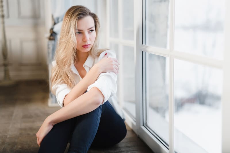 eine junge Blondine sitzt traurig am Fenster