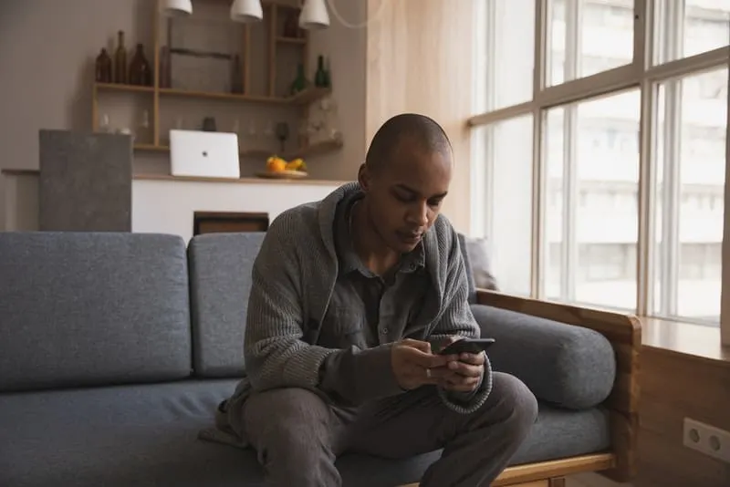 Ein Mann in einem grauen Pullover sitzt auf der Couch und schreibt eine SMS
