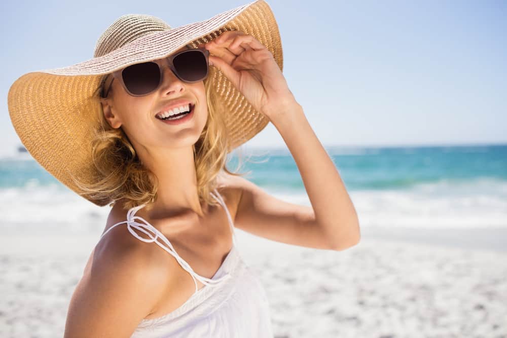 Eine lächelnde Frau mit Sonnenbrille und Hut steht am Strand