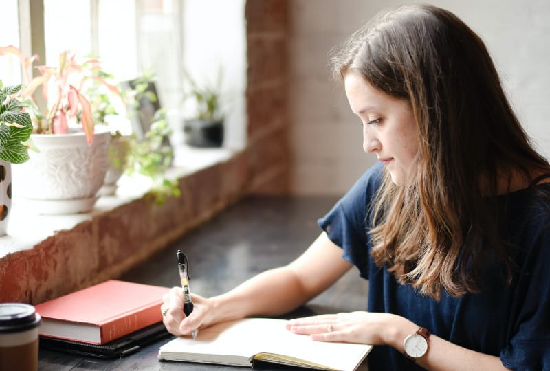 Frau, die vor schwarzem Tisch sitzt, der auf weißem Buch nahe Fenster schreibt