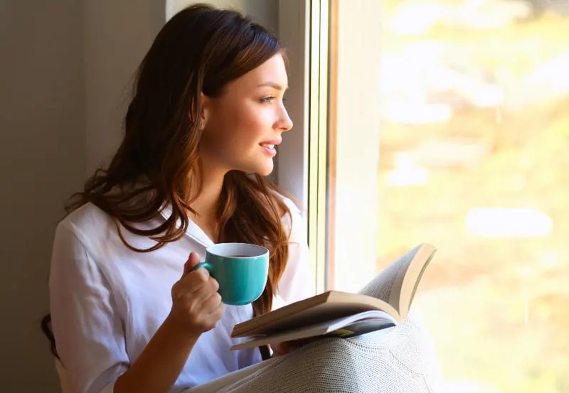 eine junge Frau, die mit einem Buch in der Hand am Fenster sitzt