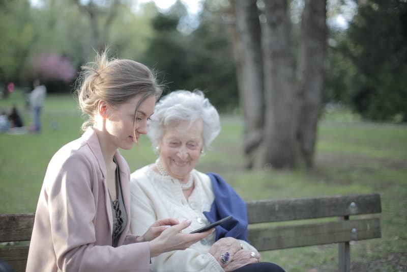 eine lächelnde ältere Frau, die auf einer Bank mit einer Frau sitzt, die eine SMS liest