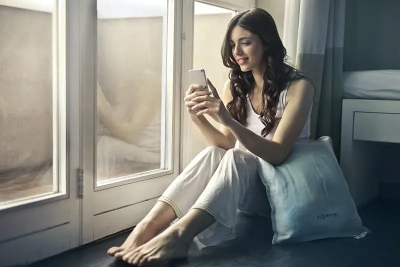 Eine Frau im Pyjama sitzt am Fenster und schreibt eine SMS