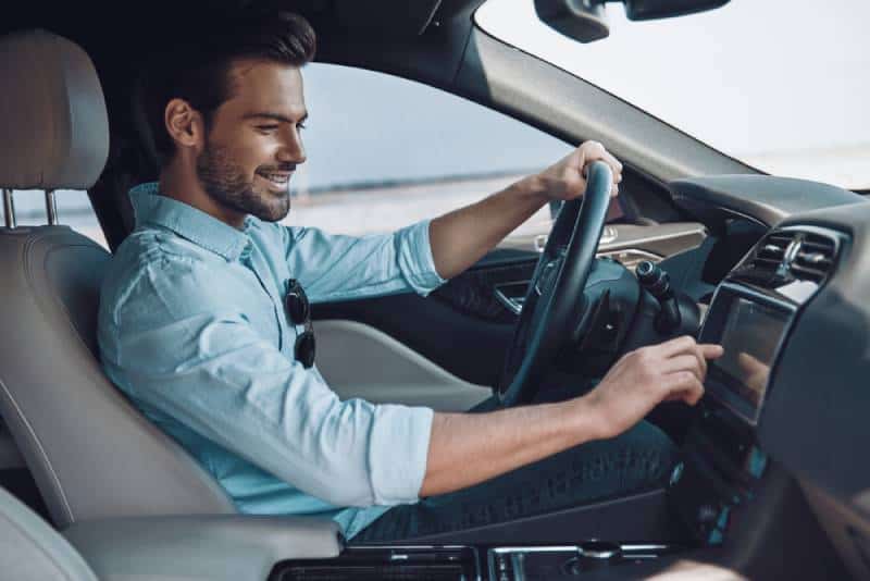 Hübscher junger Mann in der intelligenten Freizeitkleidung, die beim Fahren eines Statusautos lächelt