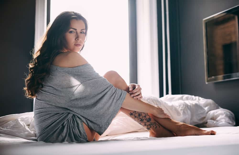 Porträt einer Brünetten mit einem Tattoo auf ihrem Bein, das auf dem Bett sitzt