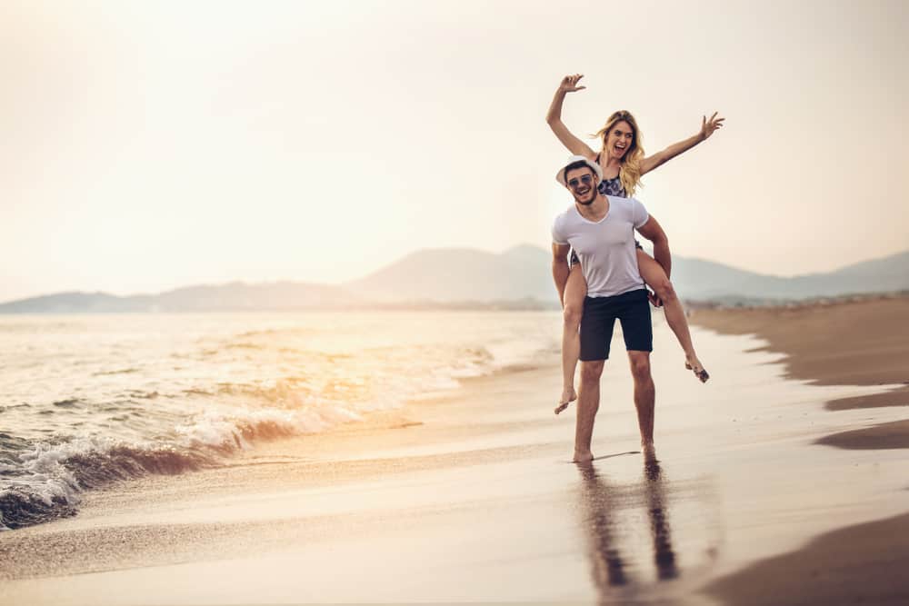 Ein Mann in Sonnenschirmen am Strand trägt seine Frau auf dem Rücken