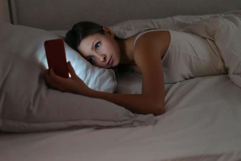Frau, die auf weißem Kissen liegt und auf ihr Telefon schaut