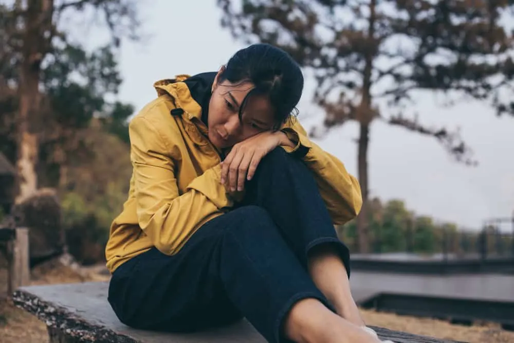 Eine asiatische Frau in einer gelben Jacke sitzt traurig auf einer Bank