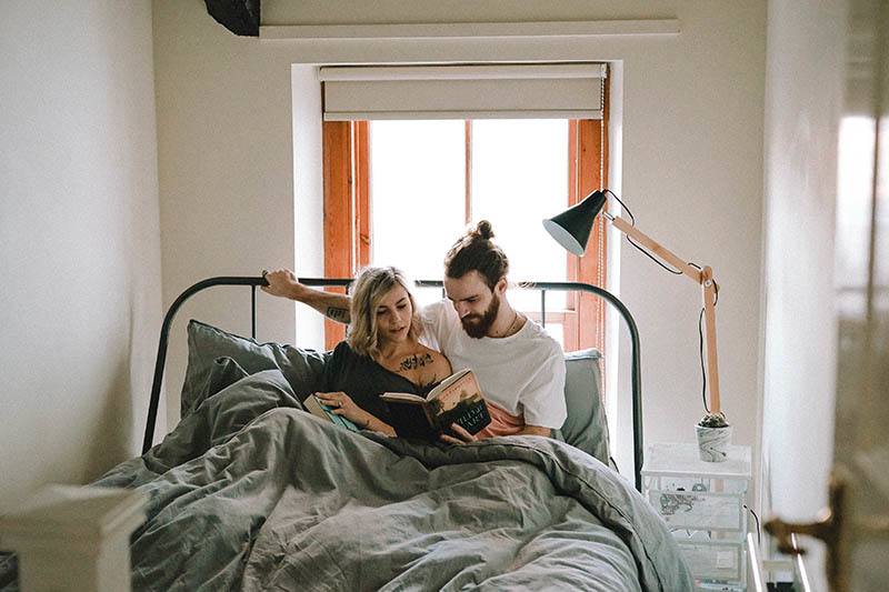 Eine Blondine mit einem Tattoo auf der Brust und ein Mann mit Bart sitzen im Bett und lesen ein Buch