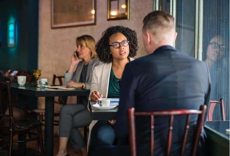 Eine schwarze Frau mit Brille und ein Mann im Anzug sitzen und reden in einem Café