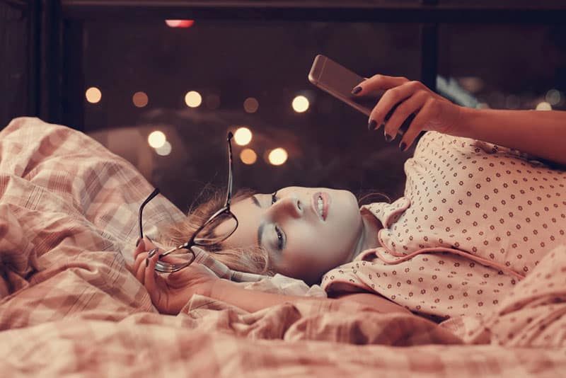 schöne junge Frau, die auf Bett liegt und auf ihr Telefon schaut