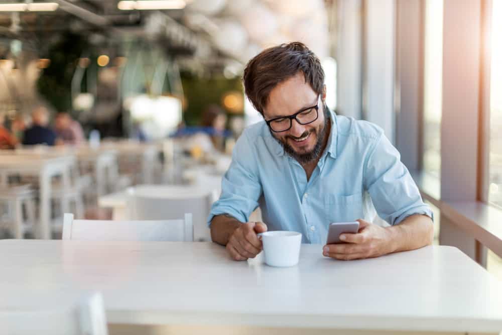 Ein lächelnder Mann mit Bart und Brille liest eine Nachricht auf seinem Handy und trinkt Kaffee