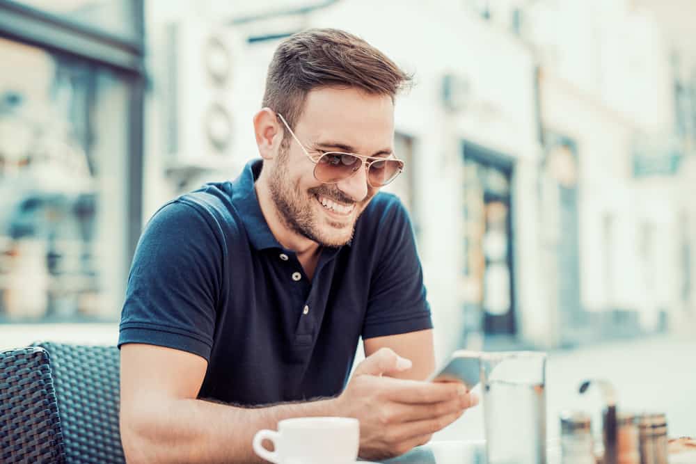 Ein lächelnder Mann mit Sonnencreme sitzt auf der Terrasse eines Cafés und benutzt ein Handy