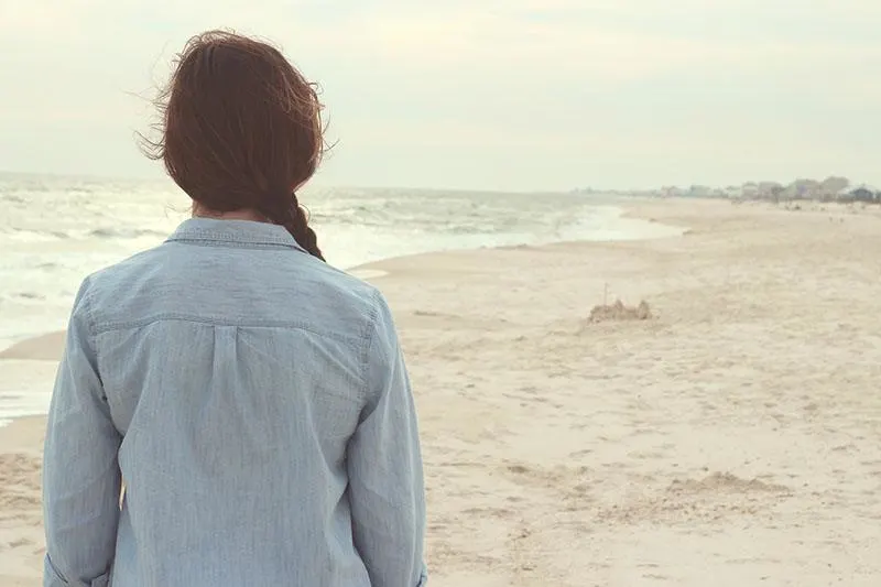 Eine Frau in Jeansjacke steht an einem Sandstrand und beobachtet das Meer