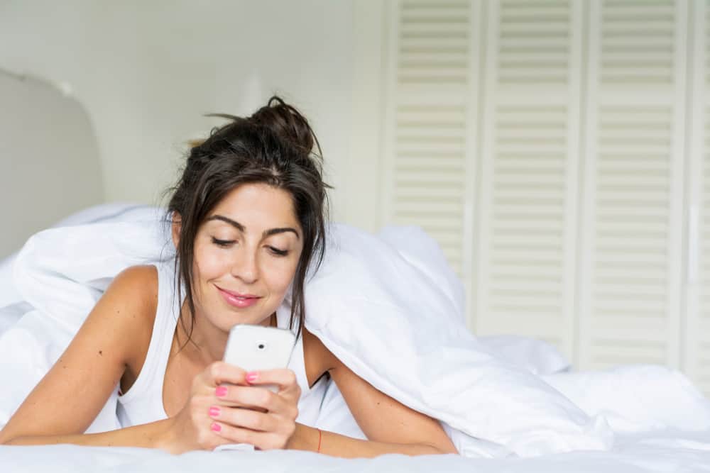 schöne Brünette, die auf dem Bett liegt und mit einem Lächeln auf ihr Handy schaut