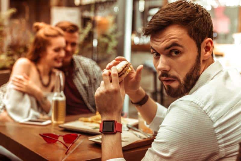 Mann mit einem Burger in den Händen, der eifersüchtig ist, während seine Freunde sich umarmen