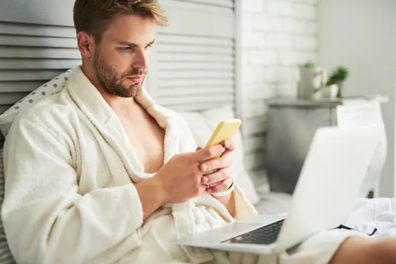 Ein Mann im Bademantel, der auf dem Bett sitzt und ein Handy benutzt