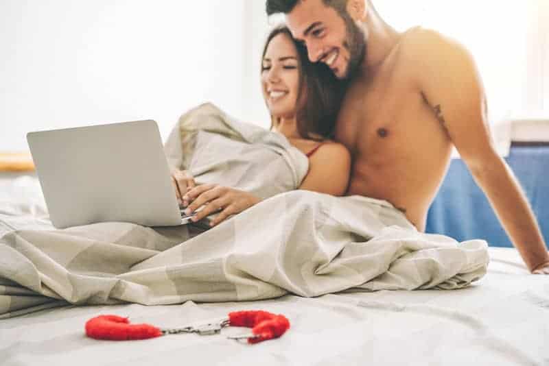 ein lächelndes Liebespaar im Bett, das etwas auf einem Laptop betrachtet