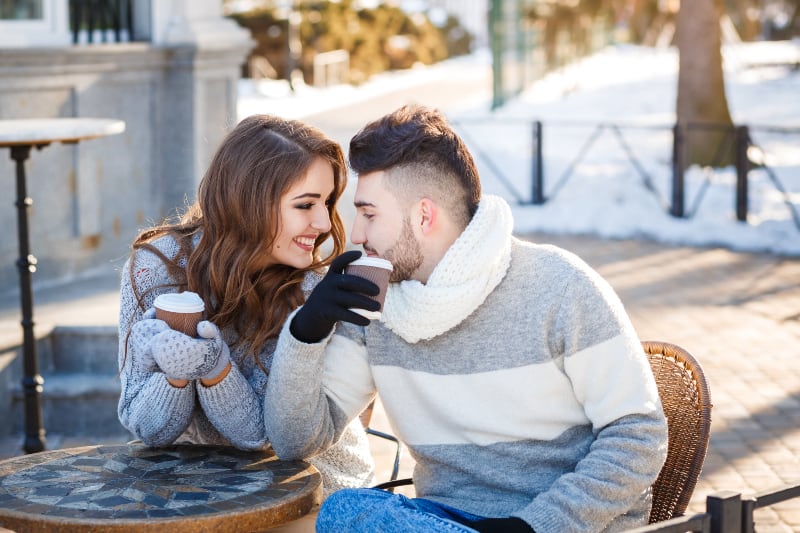 lächelndes Paar, das Sweatshirt und Handschuhe trägt, während Tasse Kaffee im Straßencafé hält