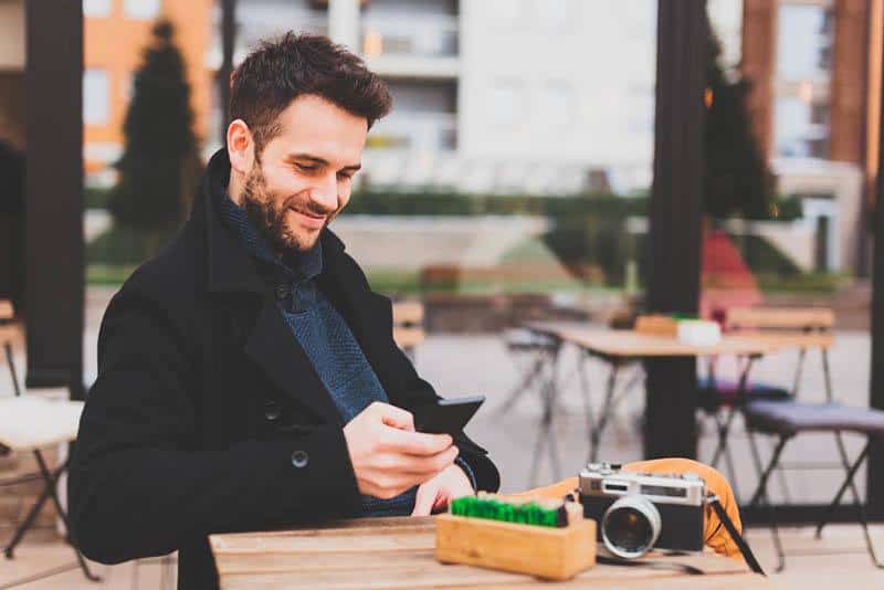 lächelnder Mann, der schwarzen Mantel trägt und auf seinem Telefon im Straßencafé tippt