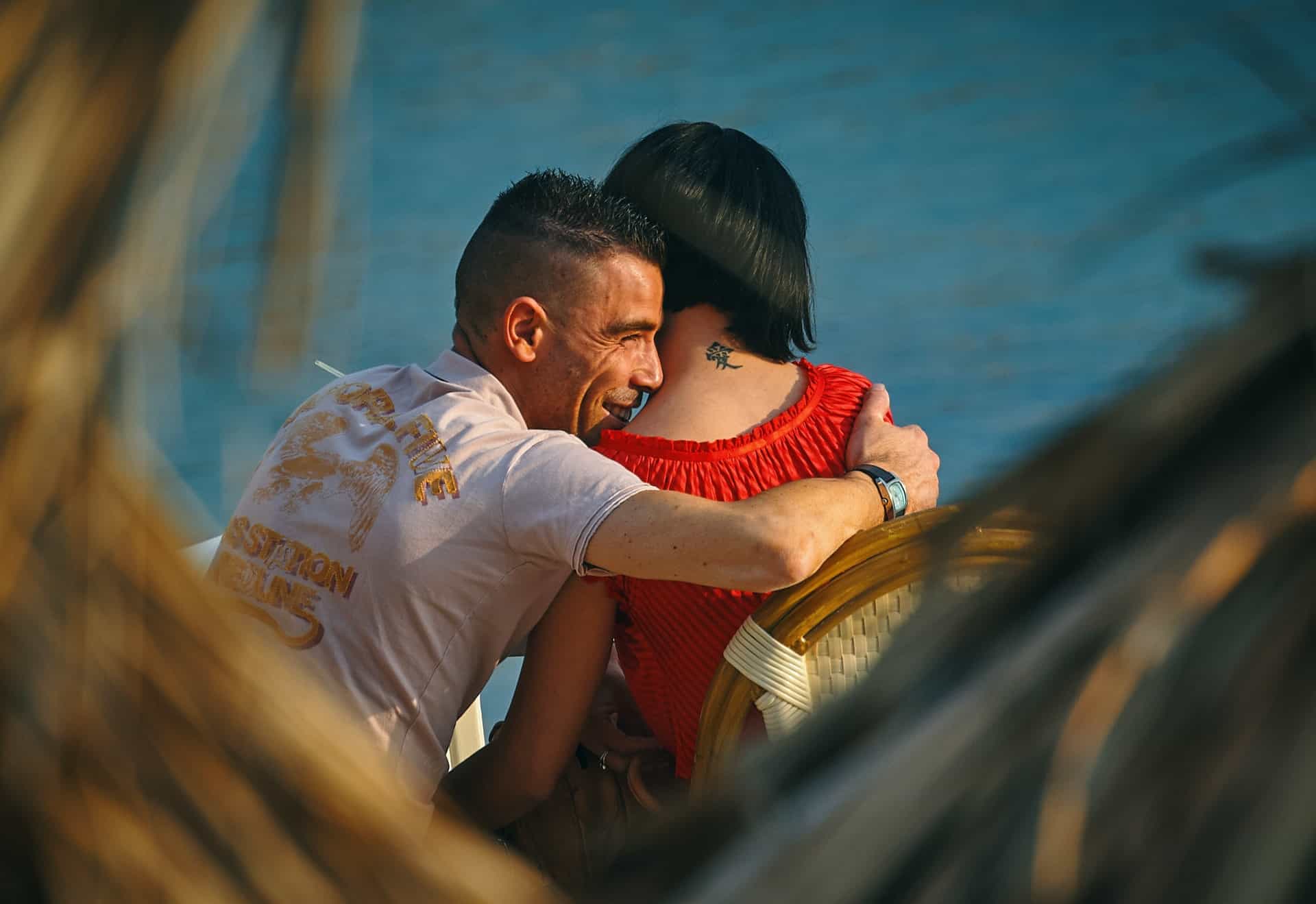 Ein lächelnder Mann umarmt eine Frau in einem roten T-Shirt, während sie am Meer sitzen
