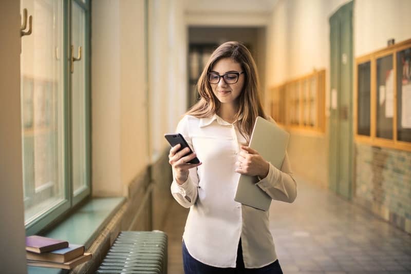 Eine glückliche Frau mit Brille steht im Flur der Schule und schreibt eine SMS