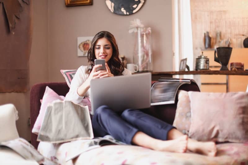 Eine lächelnde Brünette sitzt mit einem Laptop auf dem Schoß auf der Couch und schreibt eine SMS auf ihr Handy