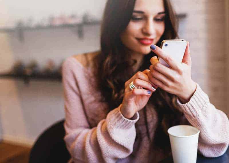 Eine lächelnde schöne Frau in einem Pullover sitzt in der Wohnung und schreibt eine SMS