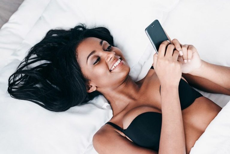 Eine schöne schwarze Frau mit einem Lächeln im Gesicht in der Unterwäsche schreibt eine Nachricht auf ihr Handy
