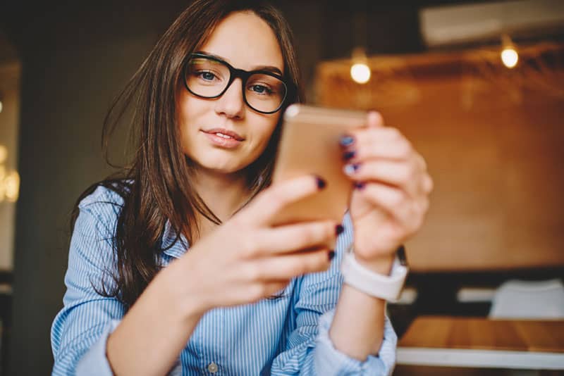 Ein schönes lächelndes Mädchen mit Brille hält ein Handy in den Händen