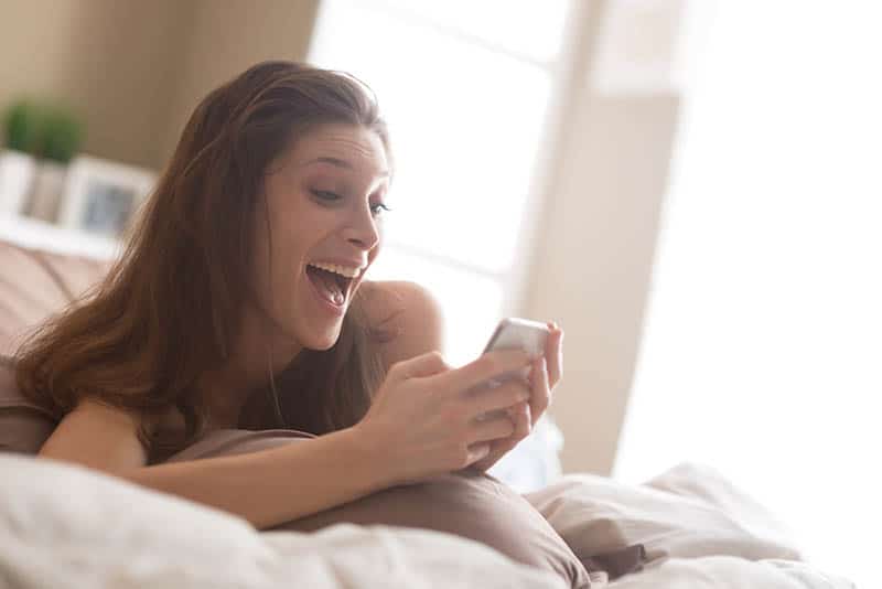 Eine überraschte lächelnde Frau liegt auf dem Bett und liest etwas auf ihrem Handy