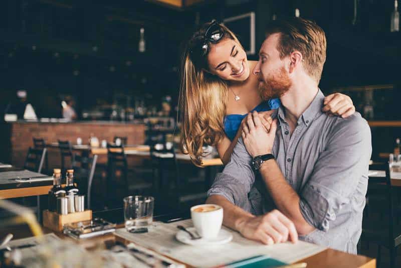 Eine Frau in einem Café umarmt einen Mann hinter ihrem Rücken