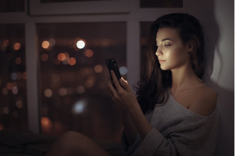 Eine attraktive Brünette sitzt abends im Haus und benutzt ein Smartphone