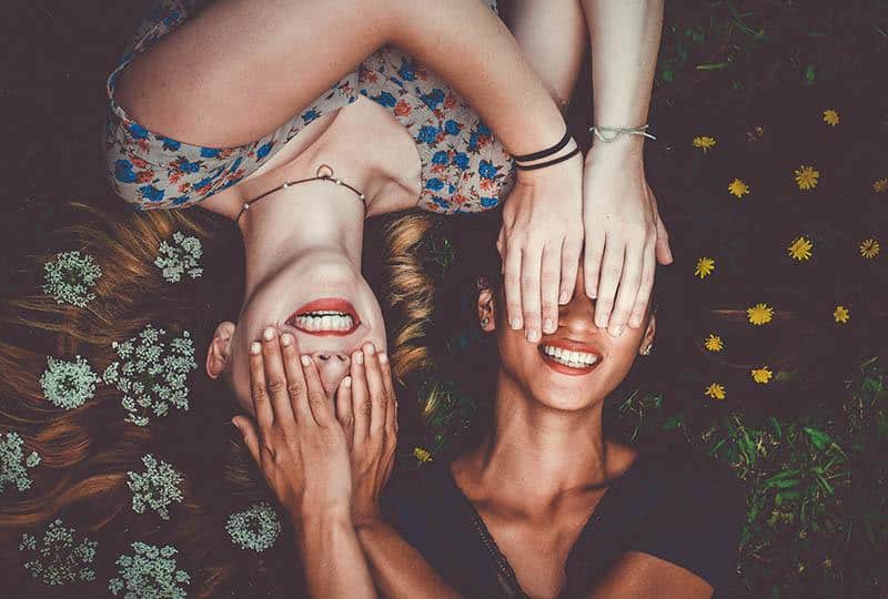 zwei lächelnde Mädchen, die im Gras liegen und ihre Hände über ihren Augen halten