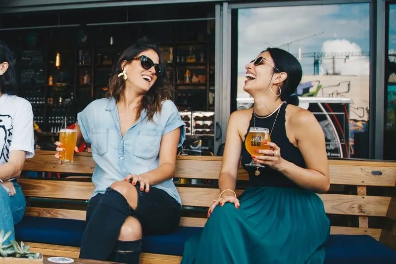 Auf der Terrasse des Cafés sitzen auf einer Holzbank zwei lächelnde Frauen, die Bier trinken
