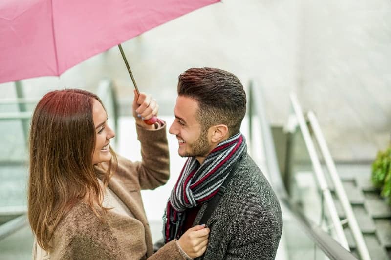 Draußen unter einem Regenschirm stehen ein lächelnder Mann und eine lächelnde Frau