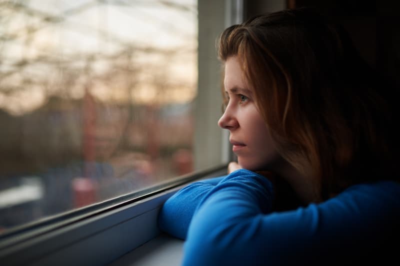 Eine traurige nachdenkliche Blondine in einem blauen T-Shirt sitzt am Fenster gelehnt und schaut nach draußen